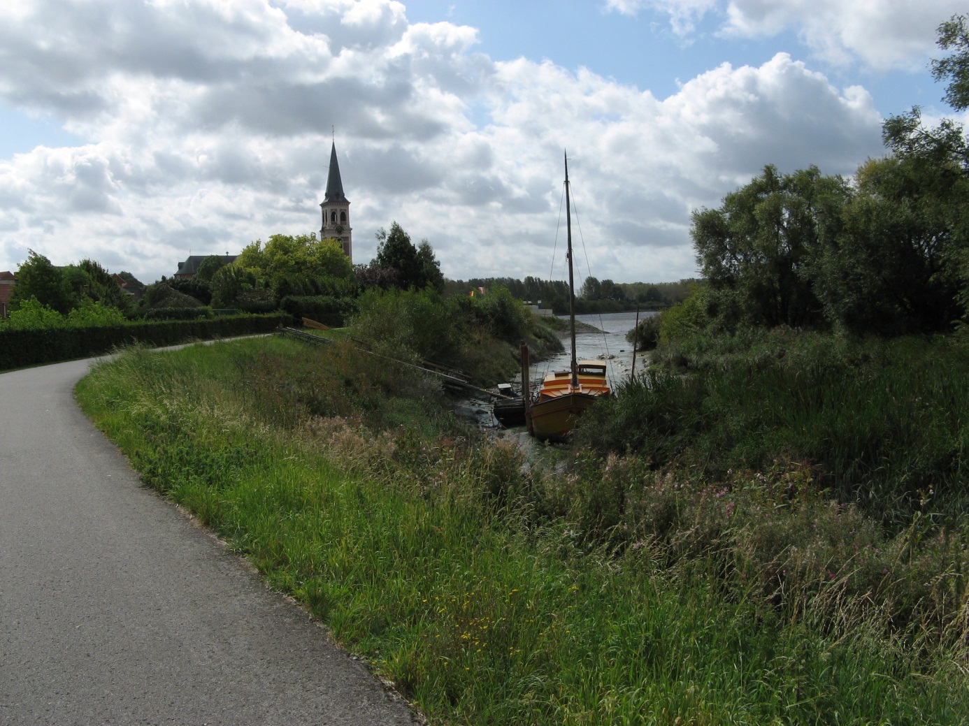 pad langs rivier met boot en een kerktoren op de achtergrond